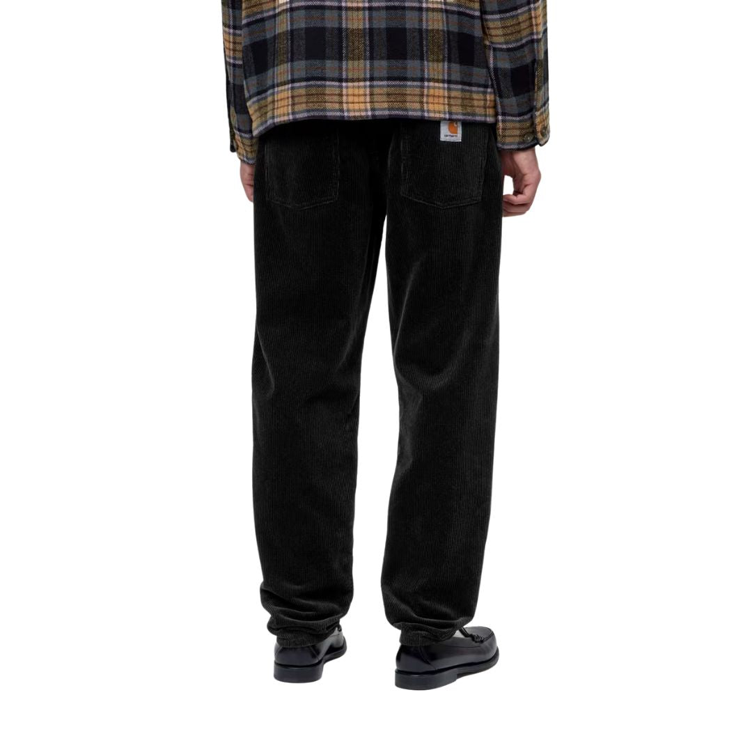 Vista trasera de un modelo con el pantalón de pana Carhartt WIP Newel Pant en color negro, camisa de cuadros y mocasines, mostrando los bolsillos traseros y la etiqueta Square Label.
