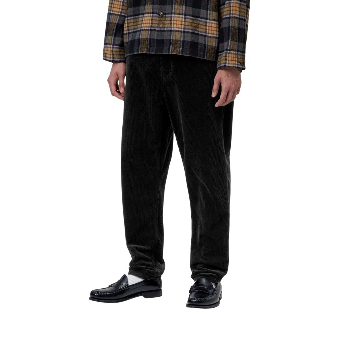 Plano medio de un modelo con el pantalón de pana Carhartt WIP Newel Pant en color negro y mocasines, visto de tres cuartos para apreciar el corte relaxed tapered en la pierna.
