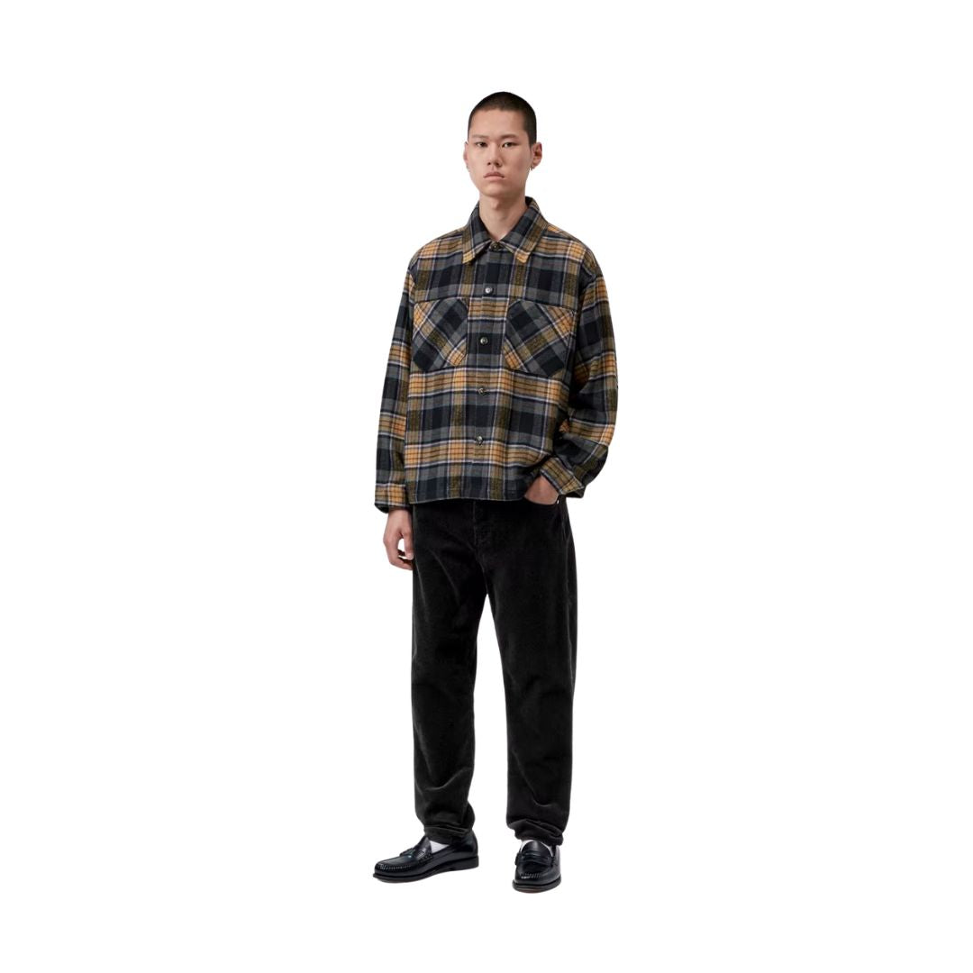 Modelo de cuerpo entero con el pantalón de pana Carhartt WIP Newel Pant en color negro, camisa de cuadros y mocasines, mostrando la caída relajada y el bajo estrecho de la pernera.