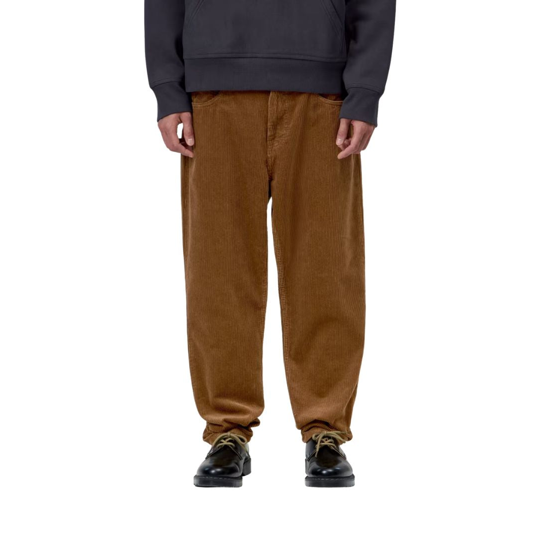 Plano frontal de un modelo con el pantalón de pana Carhartt WIP Newel Pant en color marrón cámel y sudadera con capucha, resaltando la caída recta del canalé de la pana.