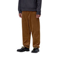 Plano medio de un modelo con el pantalón de pana Carhartt WIP Newel Pant en color marrón cámel, visto de tres cuartos para apreciar el corte relaxed tapered y el bajo con cordón.