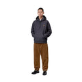 Modelo de cuerpo entero con el pantalón de pana Carhartt WIP Newel Pant en color marrón cámel y sudadera con capucha gris oscuro, mostrando la caída relajada de la pernera.