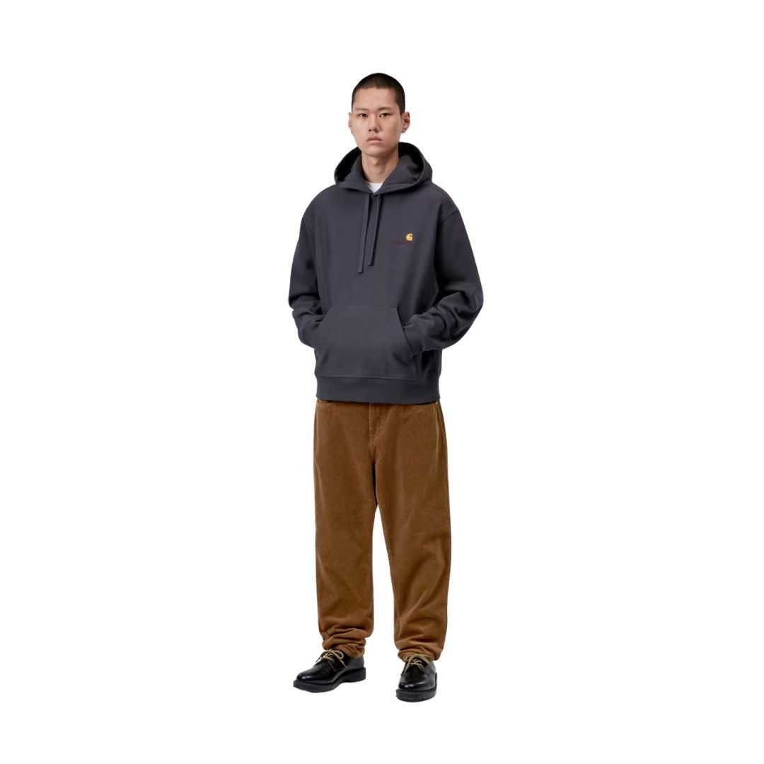 Modelo de cuerpo entero con el pantalón de pana Carhartt WIP Newel Pant en color marrón cámel y sudadera con capucha gris oscuro, mostrando la caída relajada de la pernera.
