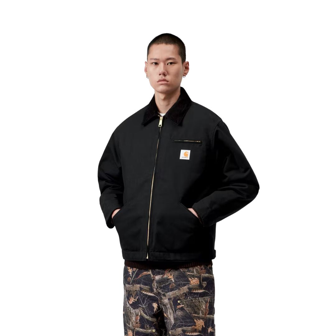 Plano medio de la Carhartt WIP OG Detroit Jacket Winter en color negro cerrada, con el modelo mostrando el cuello de pana, la cremallera dorada y la etiqueta Carhartt WIP en el pecho.
