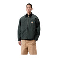 Modelo con la chaqueta acolchada Carhartt WIP OG Detroit Jacket en color Raven / Black cerrada, en plano medio, con cuello oscuro y etiqueta cuadrada en el pecho.
