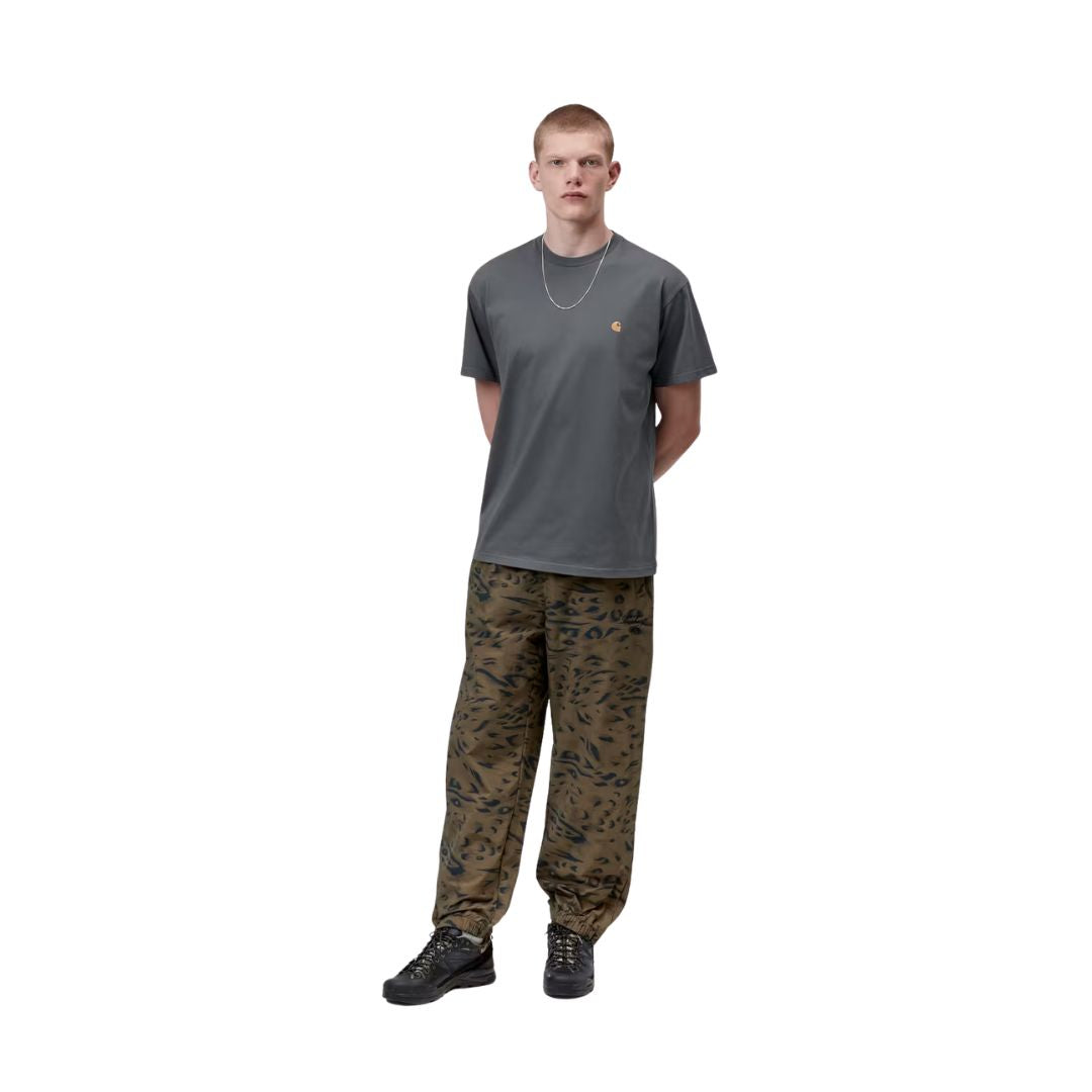 Carhartt WIP S/S Chase T-Shirt en color styx con detalle dorado, vista completa de look sobre modelo masculino. Camiseta de algodón de manga corta en gris pizarra con logotipo "C" bordado en dorado en el pecho izquierdo. Combinada con pantalón Carhartt WIP de estampado camuflaje animal print en tonos caqui y negro, y zapatillas de trekking negras.