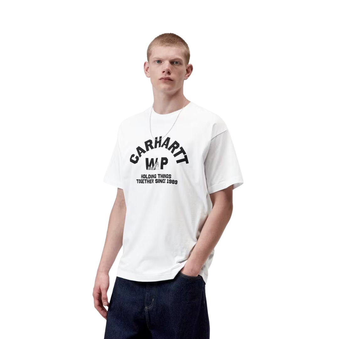 Carhartt WIP S/S Column T-Shirt en color blanco, vista frontal lateral sobre modelo masculino. Camiseta de algodón orgánico con estampado tipográfico arqueado en negro "Carhartt WIP / Holding Things Together Since 1989" en el pecho, cuello redondo y corte loose fit. Combinada con vaqueros anchos azul oscuro y cadena plateada.