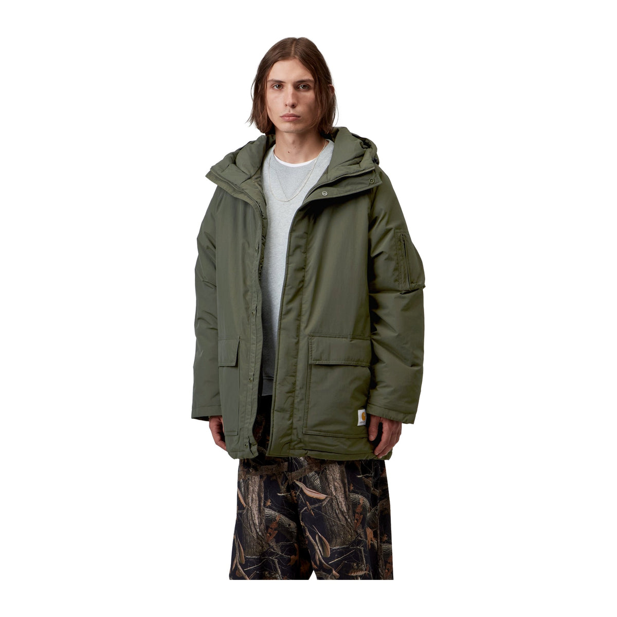 Modelo con la parka acolchada Carhartt WIP Stanwood Parka en color Cypress abierta, en plano medio, con capucha, bolsillos de parche y etiqueta cuadrada en el bajo.
