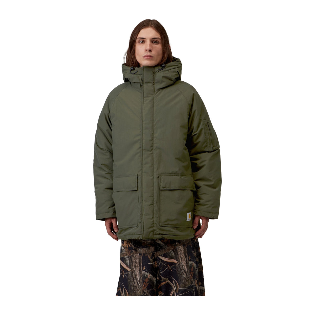 Modelo de frente con la parka Carhartt WIP Stanwood Parka en color Cypress cerrada, con la tapeta de botones, la capucha y las manos en los bolsillos de parche.