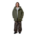 Modelo de cuerpo entero con la parka Carhartt WIP Stanwood Parka en color Cypress abierta sobre sudadera gris, con pantalón estampado y calzado negro.