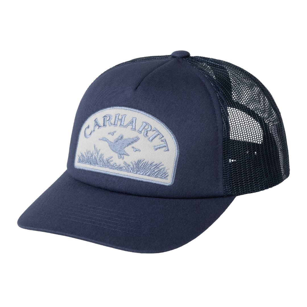 Carhartt WIP Take Off Trucker Cap en color azul marino, vista frontal de tres cuartos. Gorra trucker de sarga de algodón con panel delantero reforzado, visera curva y firme, y parche de sarga bordado con ilustración de patos volando sobre hierba y texto Carhartt en tono celeste sobre fondo blanco roto. Malla trasera en azul marino.