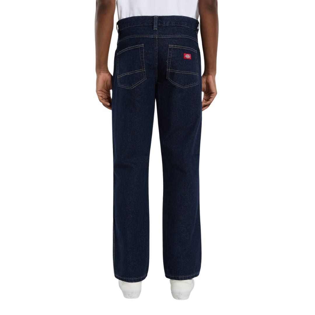 Vista trasera de modelo con vaqueros Dickies 983 Regular Straight Jeans rinsed, con dos bolsillos traseros en punta con doble costura, etiqueta roja Dickies y zapatillas blancas.
