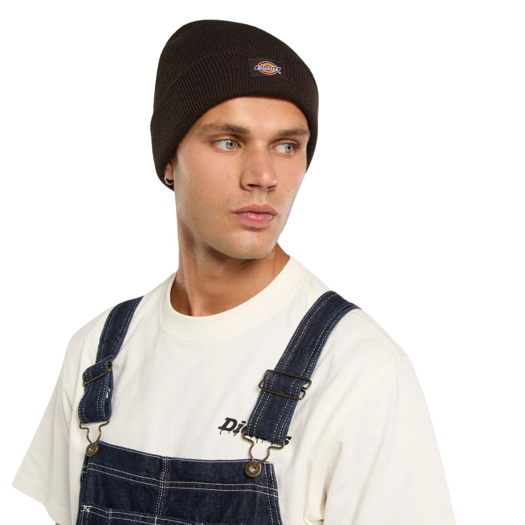 Modelo con el Dickies Gibsland Beanie marrón, gorro de punto unisex de inspiración deportiva vintage con etiqueta Dickies cosida, sobre un peto vaquero y camiseta blanca.