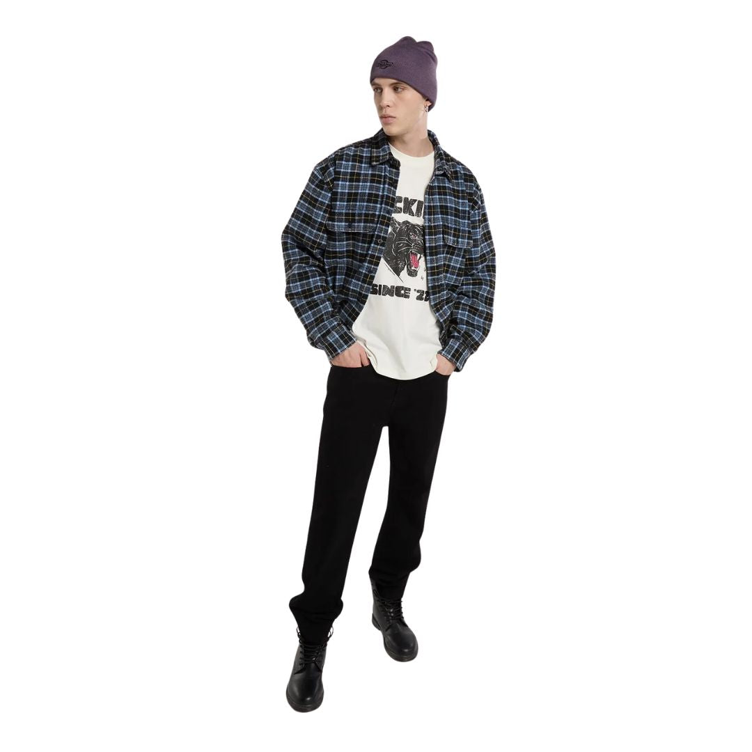 Modelo con la Dickies McDermitt SS Tee blanca crudo abierta bajo una camisa de cuadros azul, con gorro morado, pantalón negro y botas negras de caña alta.