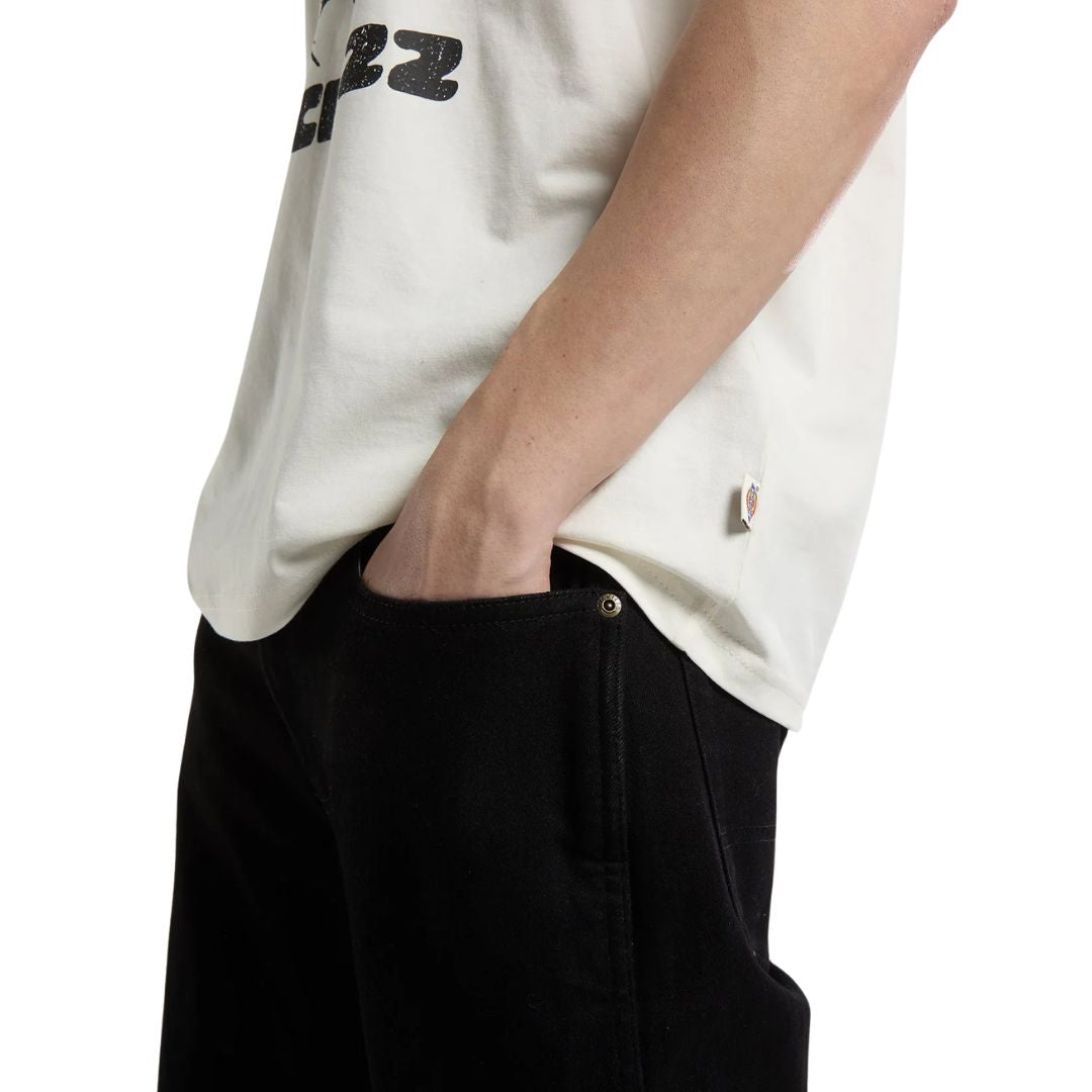 Detalle del bajo de la Dickies McDermitt SS Tee blanca crudo, con la etiqueta tejida Dickies cosida en la costura lateral, sobre un pantalón vaquero negro.