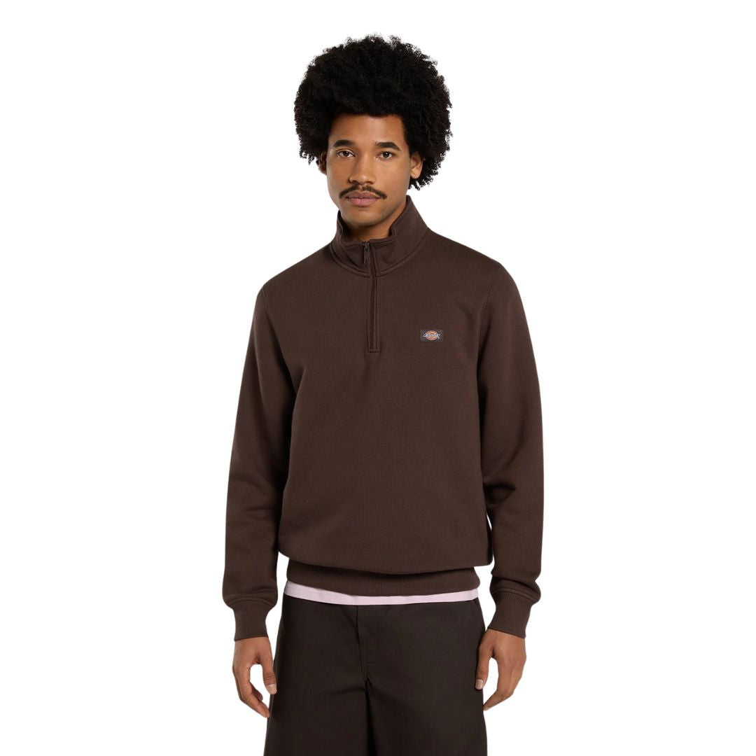 Modelo mirando a cámara con la sudadera Dickies Oakport Quarter Zip marrón chocolate de media cremallera, etiqueta Dickies en el pecho y pantalón marrón oscuro, plano frontal.