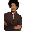 Modelo con los brazos cruzados luciendo la sudadera Dickies Oakport Quarter Zip marrón chocolate con el cuello alto y la cremallera abiertos sobre una camiseta rosa de rayas.