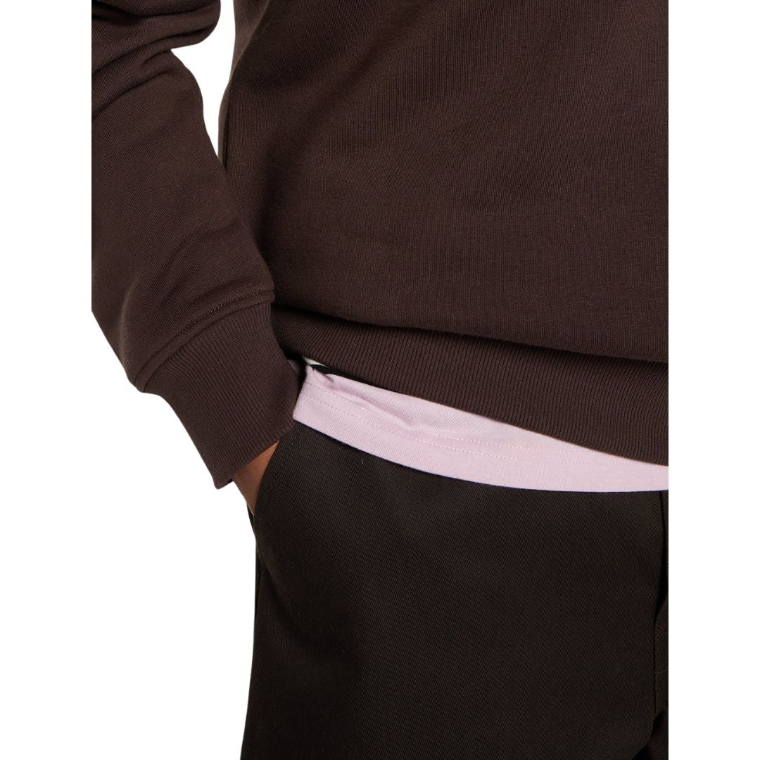 Detalle del puño y la cintura de canalé de la sudadera Dickies Oakport Quarter Zip marrón chocolate, con la mano en el bolsillo del pantalón y camiseta rosa lavanda debajo.
