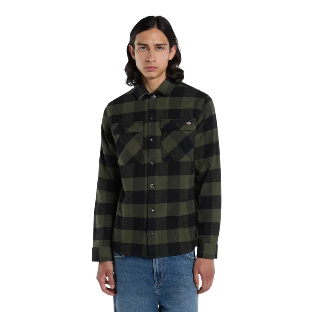 Modelo de frente con la Dickies Sacramento Shirt verde oliva y negro completamente abrochada, mostrando los dos bolsillos en el pecho, sobre pantalón vaquero azul.