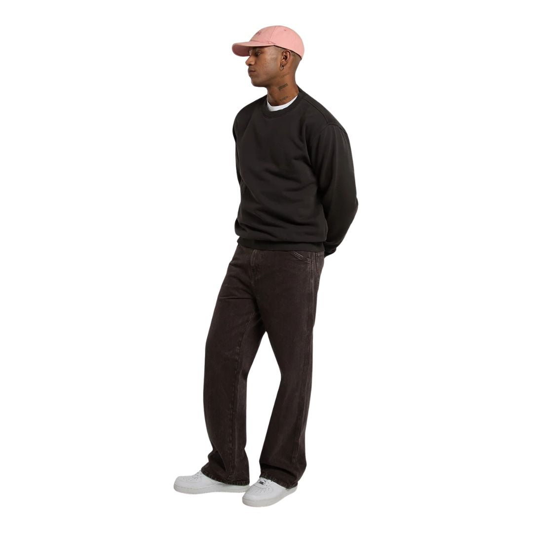 Modelo con la Dickies Sutcliffe Sweatshirt marrón oscuro, gorra rosa, pantalón vaquero marrón oscuro y zapatillas blancas, en un plano de cuerpo entero de perfil.