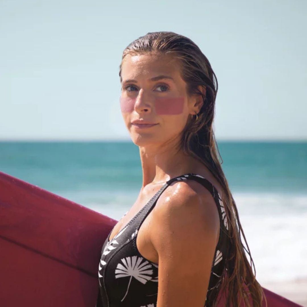 Mujer surfista con protector solar rosa en las mejillas usando stick solar SPF50+ bio resistente al agua en la playa.