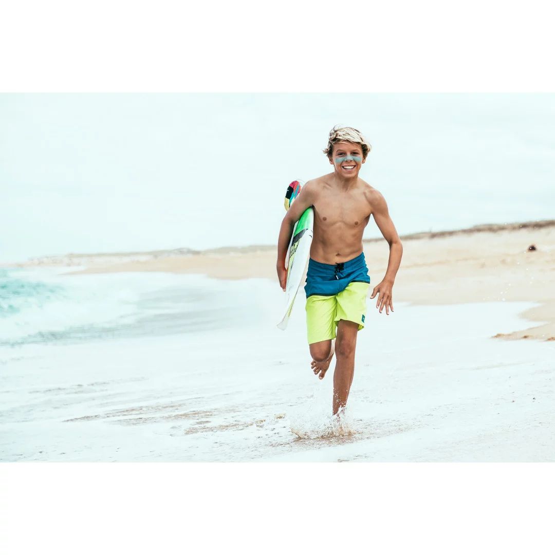 Niño surfista corriendo por la playa con protector solar turquesa en la cara usando stick SPF50+ resistente al agua.