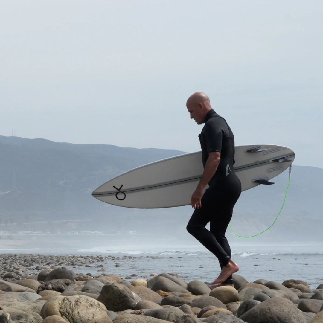Kelly Slater caminando por rocas junto al mar llevando su tabla bajo el brazo con el set de quillas Firewire Endorfins KSR instalado en los cajetines Futures.