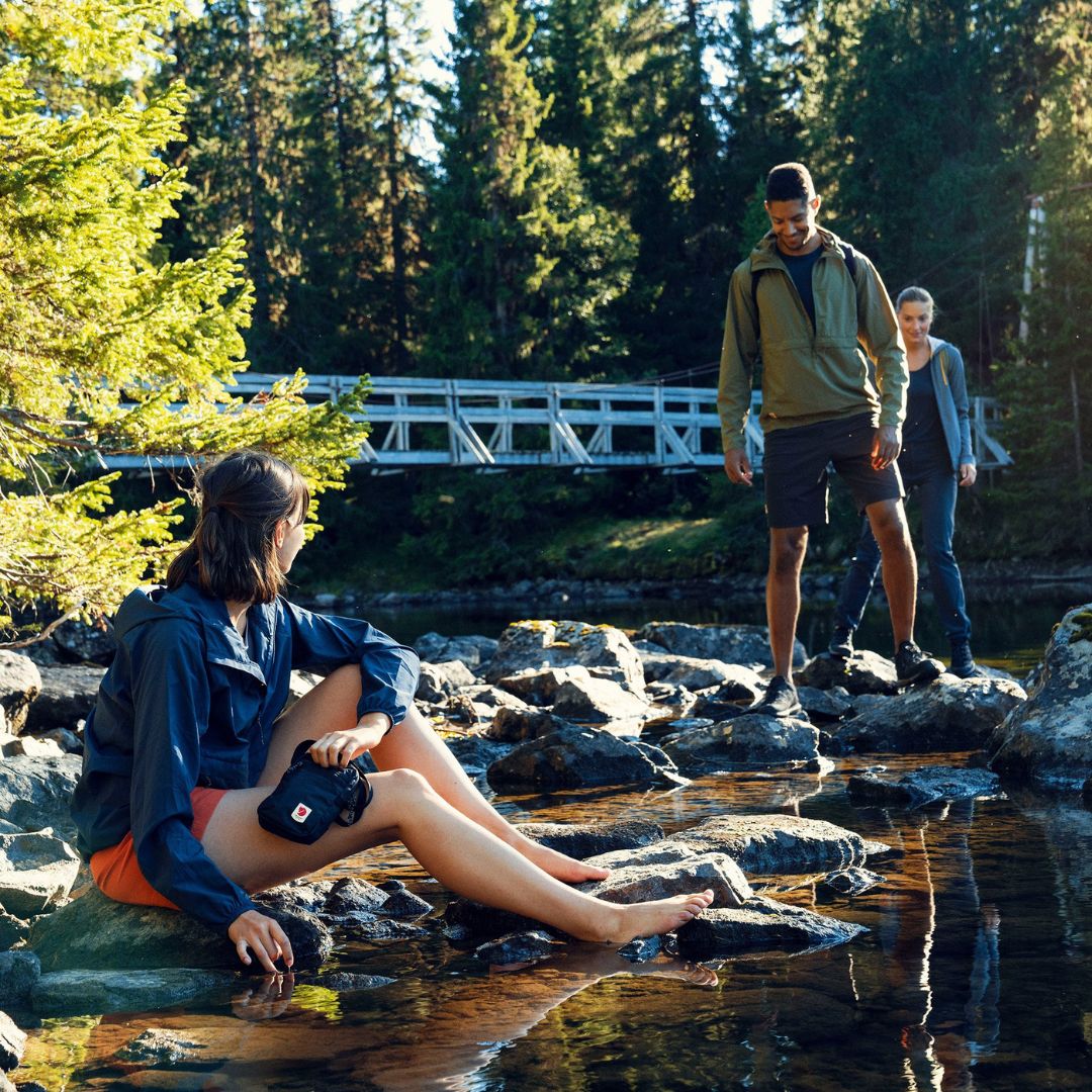 Escena de senderismo junto a un río donde una mujer sentada en las rocas sujeta la bandolera Fjällräven High Coast Pocket mientras otros excursionistas se aproximan.