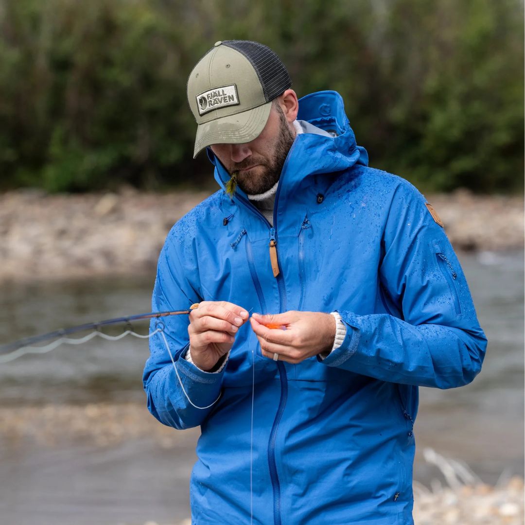 Hombre con gorra Fjällräven Långtradarkeps ajustando equipo de pesca en entorno natural.