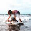 Adulto enseñando a una niña a surfear con la tabla mini long Flysurf Cold Hawaii 8.0 en la orilla.