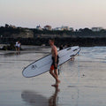 Surfista caminando por la playa con la tabla mini long Flysurf Cold Hawaii 8.0 y leash azul.