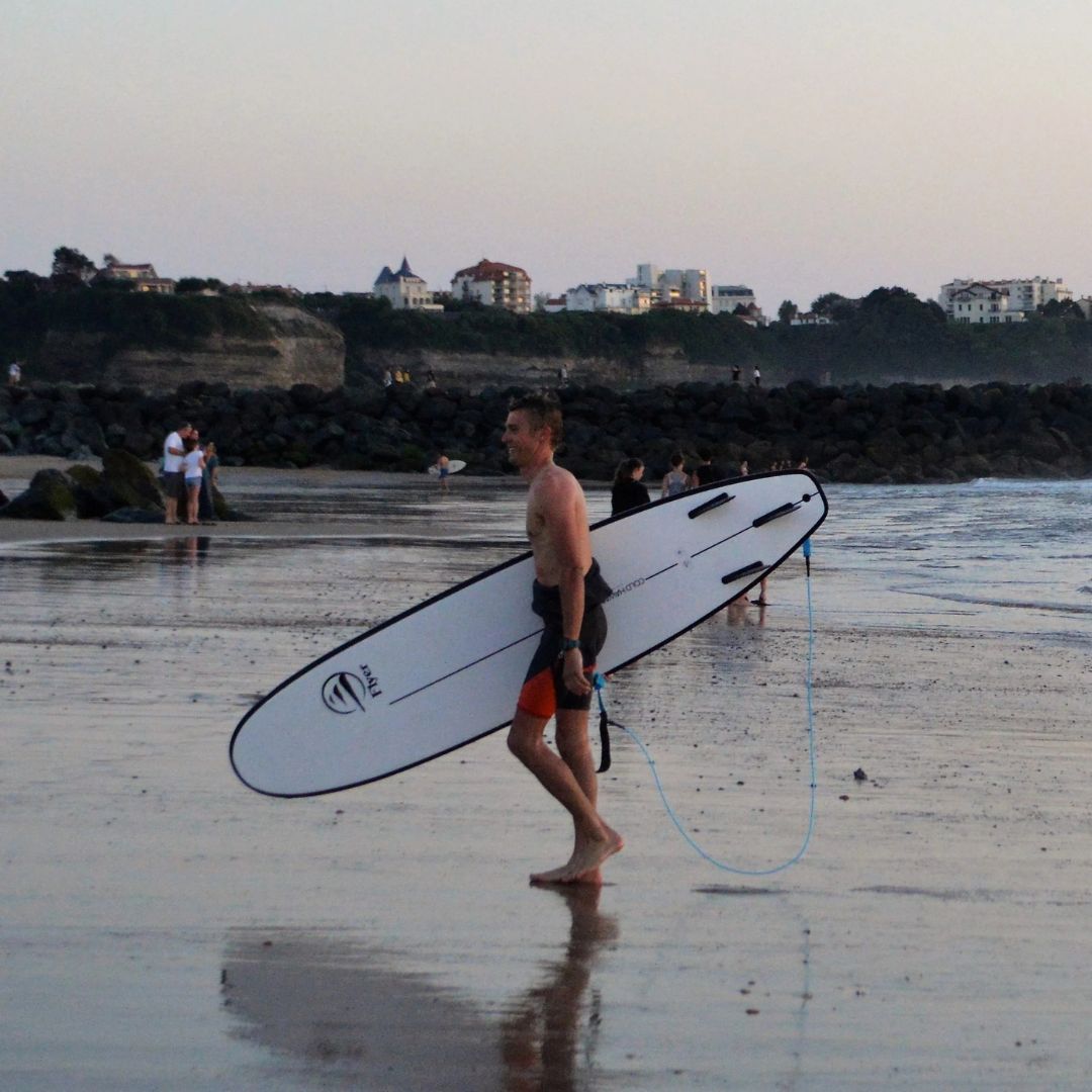 Surfista caminando por la playa con la tabla mini long Flysurf Cold Hawaii 8.0 y leash azul.