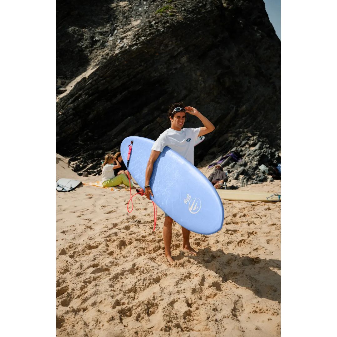 Surfista caminando por la playa con tabla de surf softboard FLYSURF Do It Your Wave 6.4 Blue Wave azul, tabla evolutiva con volumen para facilitar la remada.