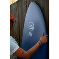 Detalle de la tabla de surf FLYSURF Do It Your Wave Blue Wave color azul apoyada verticalmente mostrando el deck con logo Fly y shape compacto.