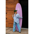 Surfista transportando la tabla de surf Flysurf Sea Calling 7.2 rosa junto a una pared de madera.
