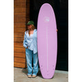 Tabla de surf rosa Flysurf Sea Calling 7.2 apoyada verticalmente junto a surfista mostrando tamaño completo.