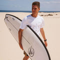 Surfista caminando por la playa con la tabla de surf evolutiva Flysurf The Traveller 6.6 blanca bajo el brazo.