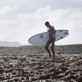Surfista caminando sobre rocas con la tabla evolutiva Flysurf The Traveller 6.6 blanca diseñada para progresar en el surf.