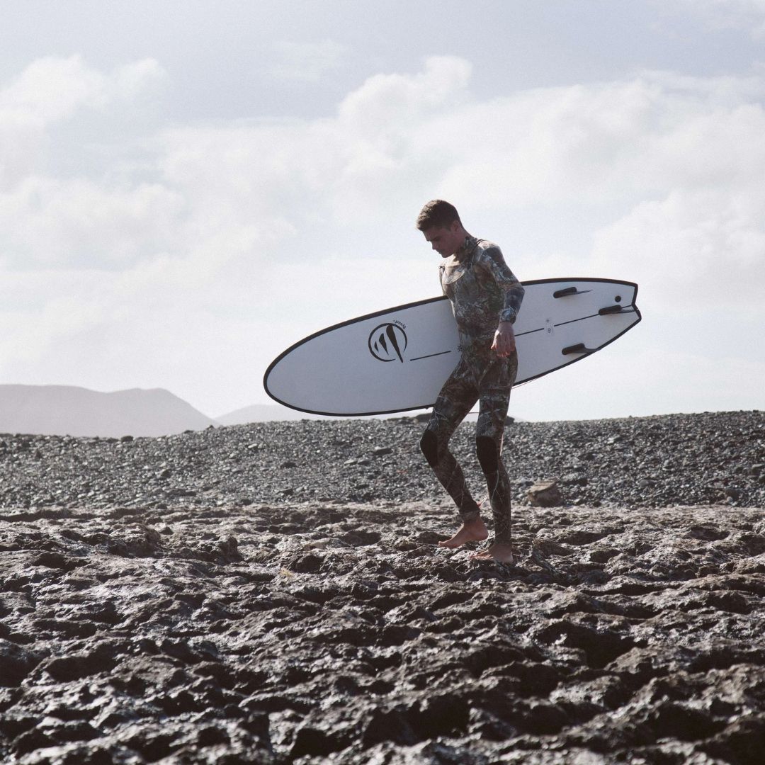 Surfista caminando sobre rocas con la tabla evolutiva Flysurf The Traveller 6.6 blanca diseñada para progresar en el surf.