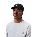 Carhartt WIP Madison Logo Cap en color negro con detalle blanco, vista frontal sobre modelo masculino. Gorra de seis paneles sin estructura en sarga prelavada negra con logo duck bordado en blanco, visera curva y hebilla metálica ajustable. Combinada con sudadera gris de cuello redondo con logo American Script de Carhartt WIP y cadena plateada.
