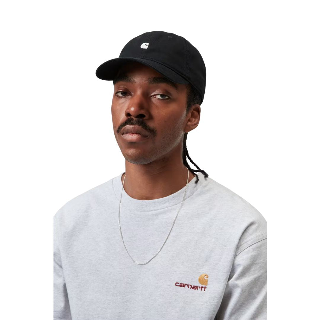 Carhartt WIP Madison Logo Cap en color negro con detalle blanco, vista frontal sobre modelo masculino. Gorra de seis paneles sin estructura en sarga prelavada negra con logo duck bordado en blanco, visera curva y hebilla metálica ajustable. Combinada con sudadera gris de cuello redondo con logo American Script de Carhartt WIP y cadena plateada.