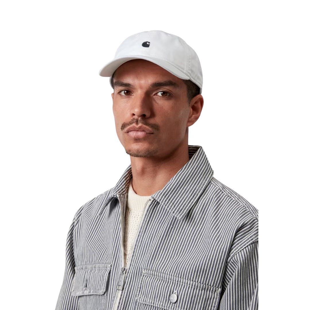 Carhartt WIP Madison Logo Cap en color blanco con detalle negro sobre modelo masculino, vista frontal. Gorra de seis paneles sin estructura en sarga prelavada blanca con logo duck bordado en negro, visera curva y hebilla metálica ajustable. Combinada con chaqueta workwear Carhartt WIP de rayas y jersey crema.