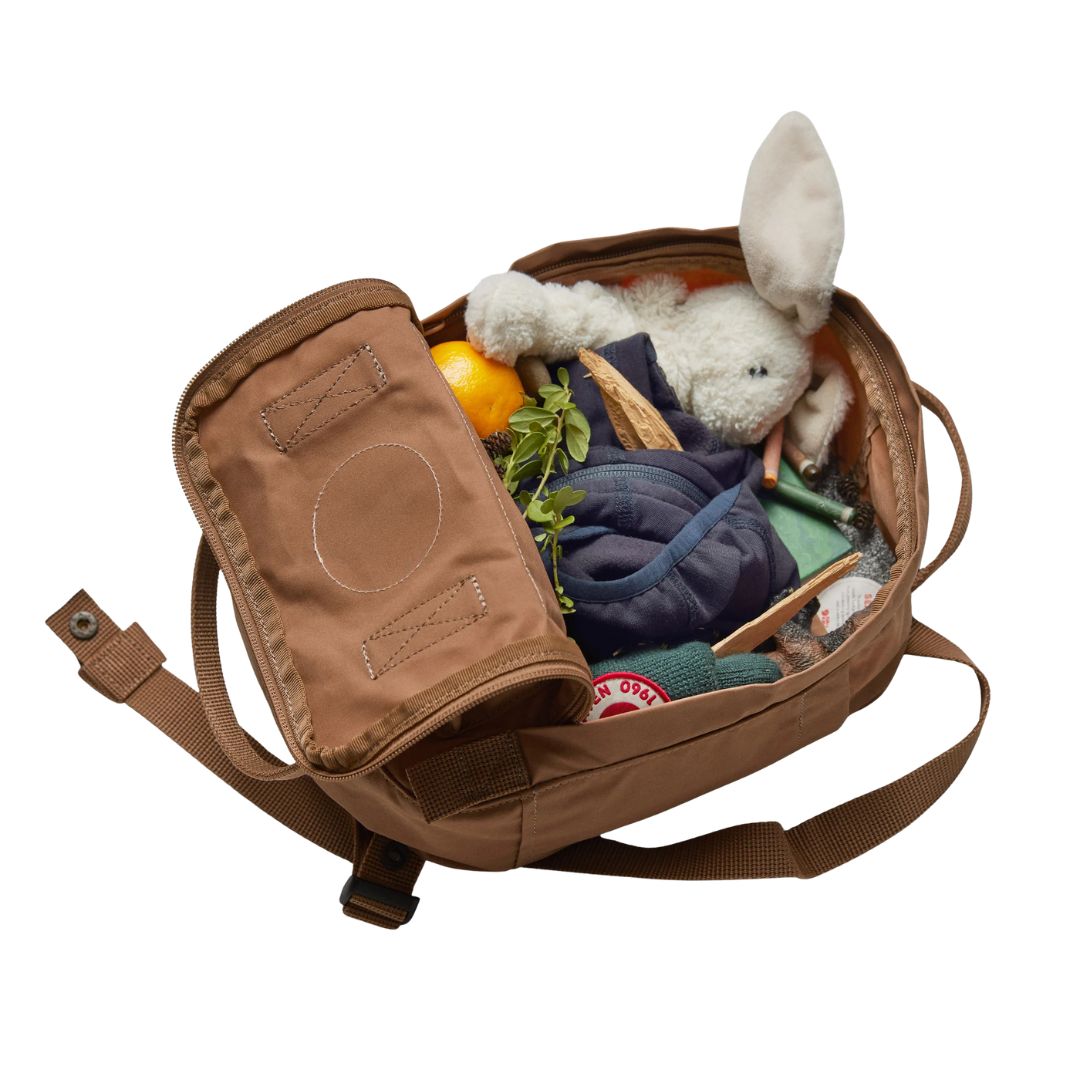 Mochila Fjällräven Kånken Mini abierta mostrando su interior con juguetes, ropa infantil y merienda, en color arena