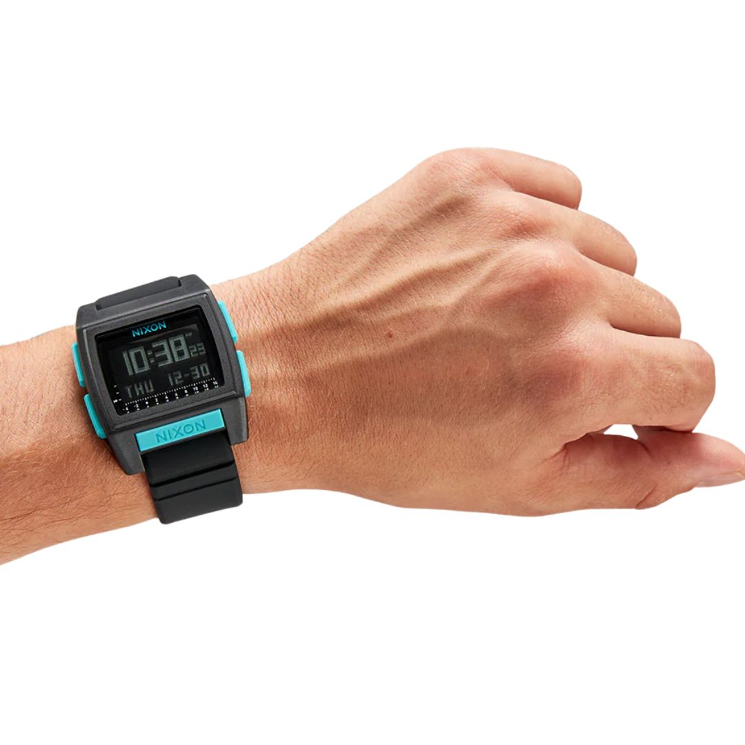 Persona usando el reloj Nixon Base Tide Pro negro con detalles azul turquesa sobre una tabla de surf.