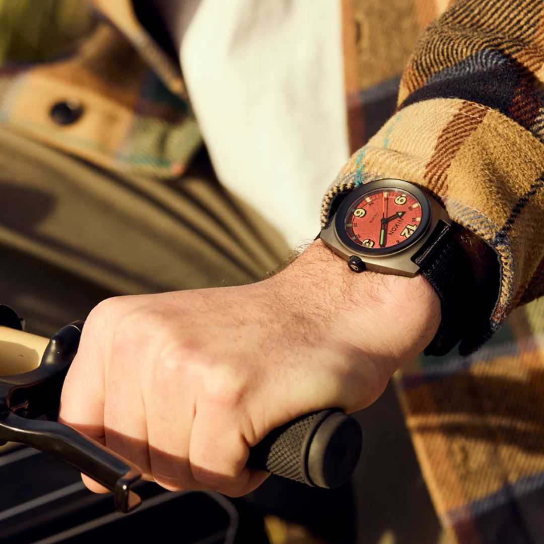 Reloj Nixon Clutch 42 Matte Gunmetal Hot Sauce en la muñeca de un usuario sujetando el manillar de una bicicleta bajo la luz del atardecer.