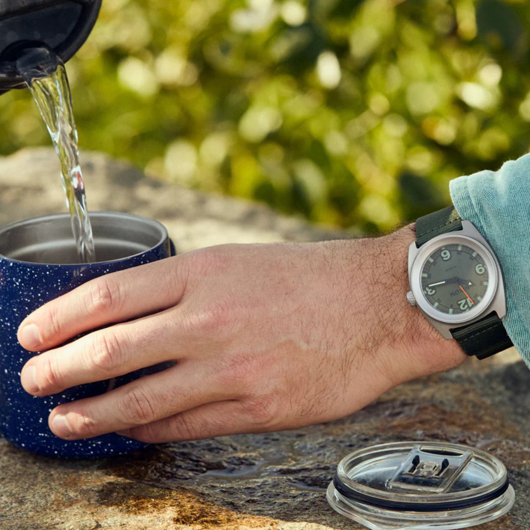 Reloj Nixon Clutch 42 Matte Silver Thyme en la muñeca de un usuario al aire libre vertiendo café, mostrando su diseño aventurero.