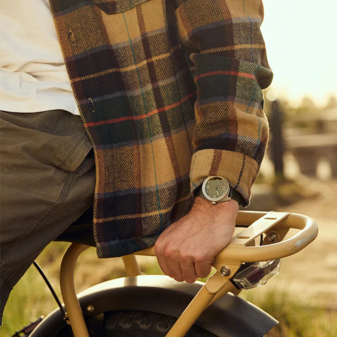 Reloj Nixon Clutch 42 Matte Silver Thyme en la muñeca de un hombre con camisa de cuadros sujetando una bicicleta bajo la luz del atardecer.