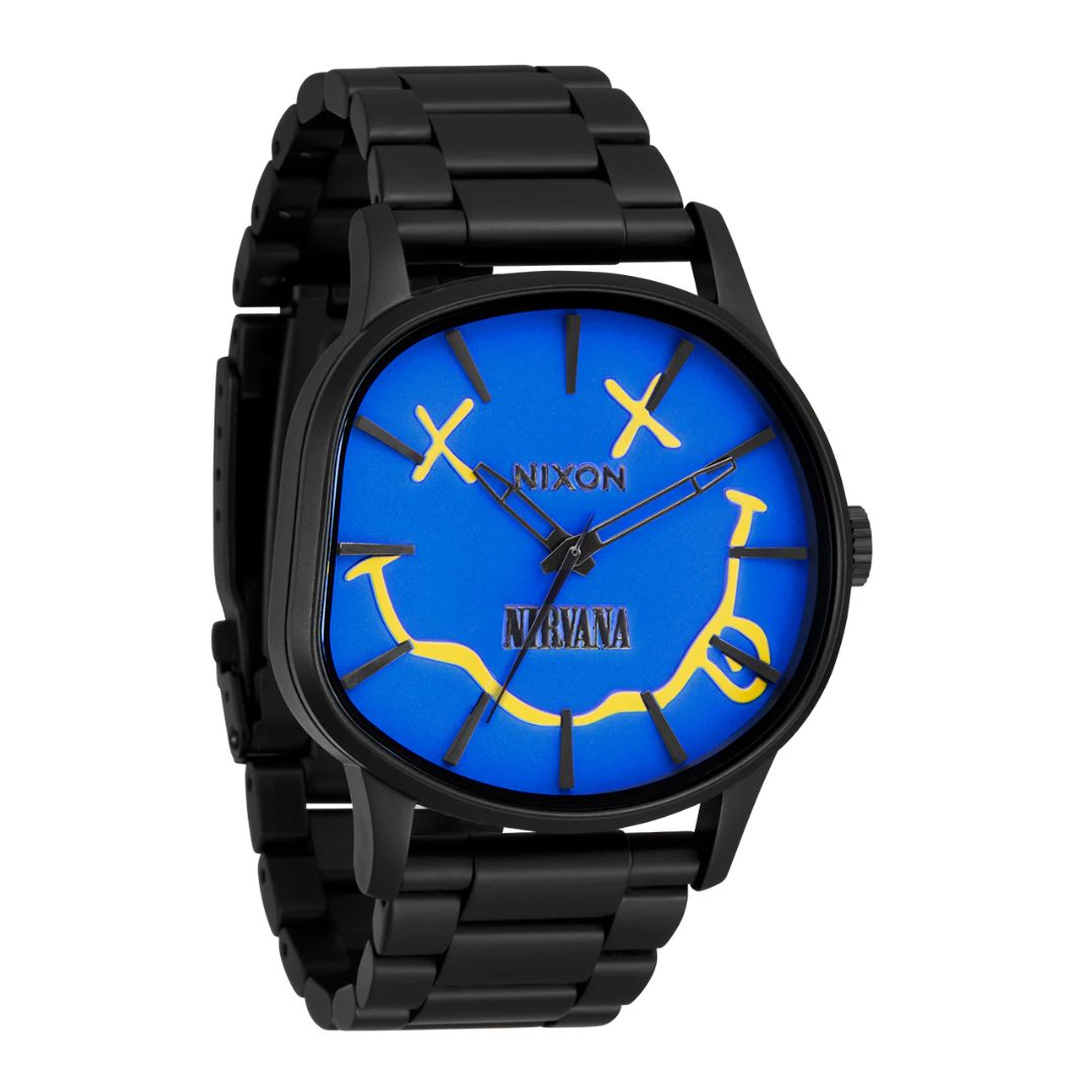 Reloj Nixon Nirvana Sentry Wobble Yellow con esfera azul y diseño del smiley amarillo de Nirvana, caja negra y correa metálica.