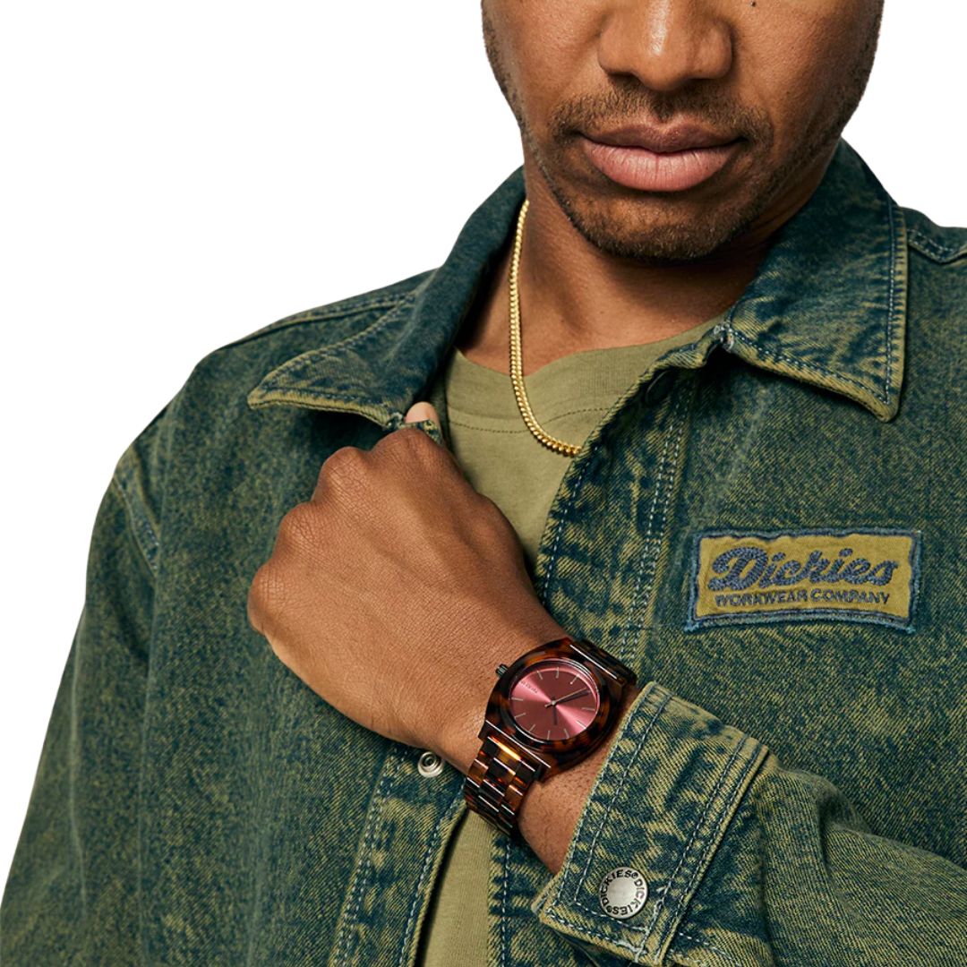 Hombre con chaqueta Dickies mostrando el reloj Nixon Time Teller Acetate tortoise oxblood.
