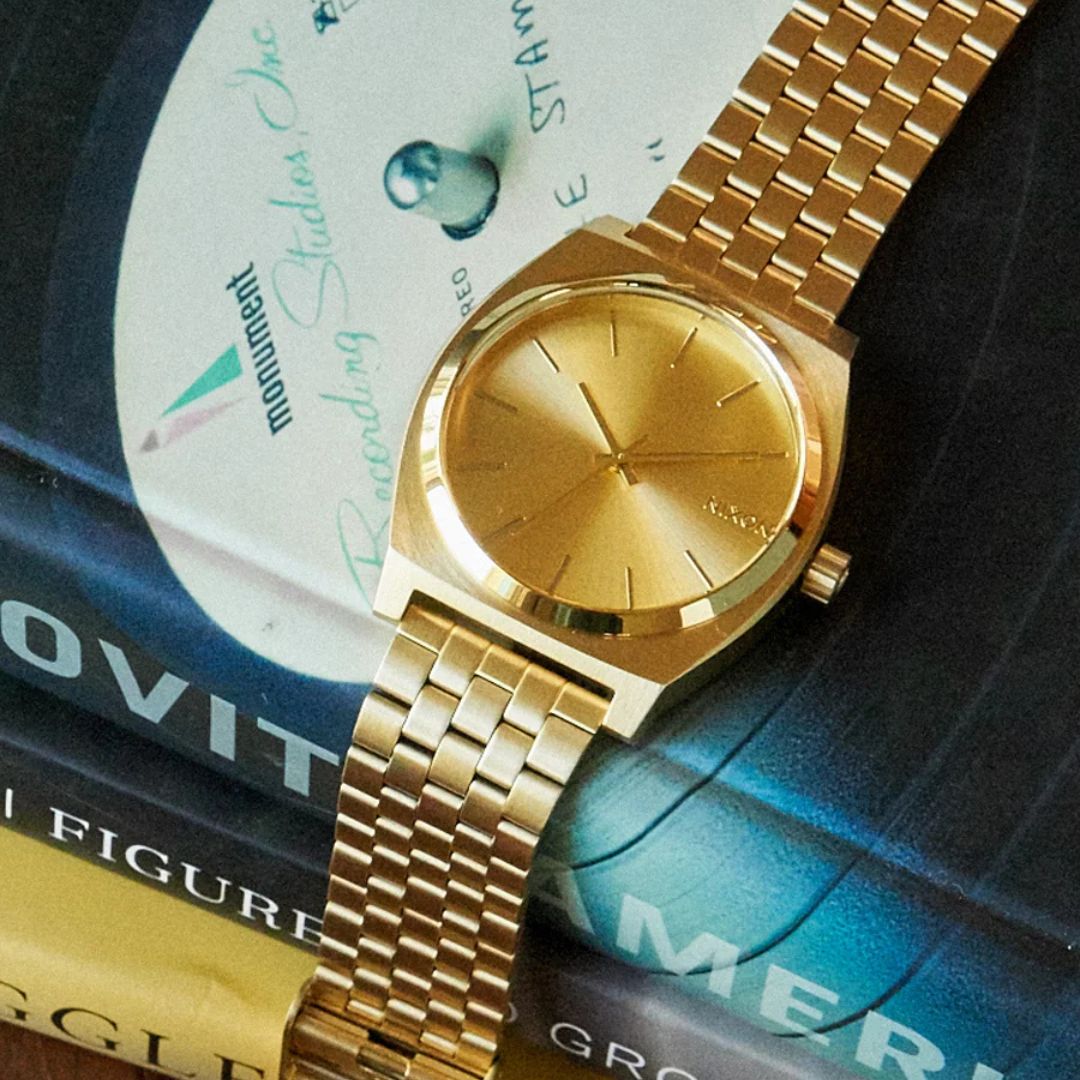 Reloj Nixon Time Teller All Gold sobre vinilos y libros, destacando su acabado dorado brillante.